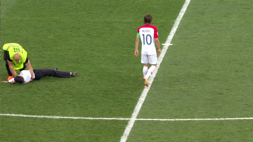 Auf einem Fussballfeld ist mittig ein Spieler in weißem Trikot von hinten zu sehen. Links wird eine liegende Person von einem Ordner festgehalten.