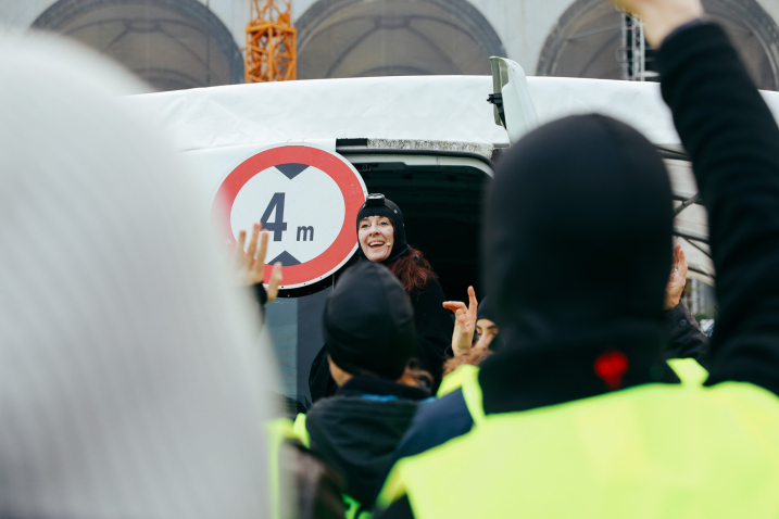 Eine Frau steht lächelnd in der geöffneten Tür eines Fahrzeugs mit einem 4-Meter-Verkehrsschild, während mehrere Personen in Warnwesten vor ihr die Hände heben.