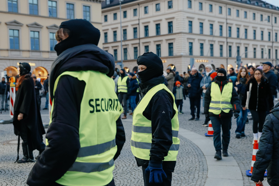 Einige vermummte und mit Sicherheitswesten bekleidete Personen stehen auf einem Platz vor einer Menschenmenge und historischen Gebäuden im Hintergrund.