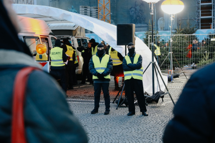 In front of and under a white tent roof, there are several masked people wearing safety vests. Under the tent roof, there are two white vans with their doors open.