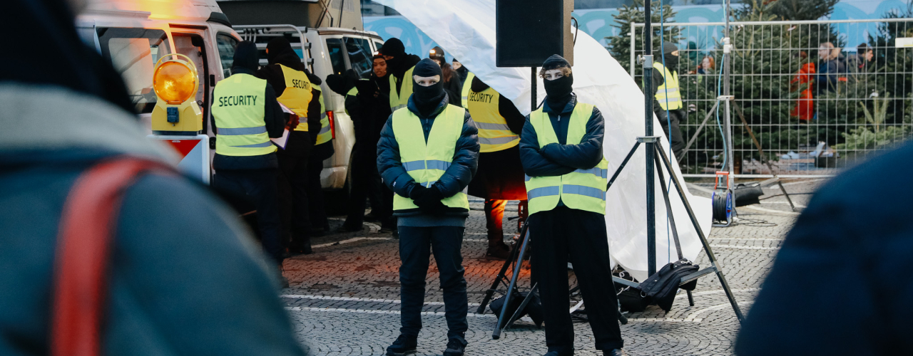 Vor und unter einem weißen Zeltdach stehen mehrere vermummte Personen mit Sicherheitswesten, unter dem Zeltdach stehen zwei weiße Transporter mit geöffneten Türen.