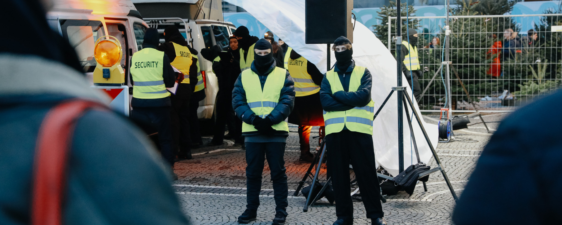 Vor und unter einem weißen Zeltdach stehen mehrere vermummte Personen mit Sicherheitswesten, unter dem Zeltdach stehen zwei weiße Transporter mit geöffneten Türen.