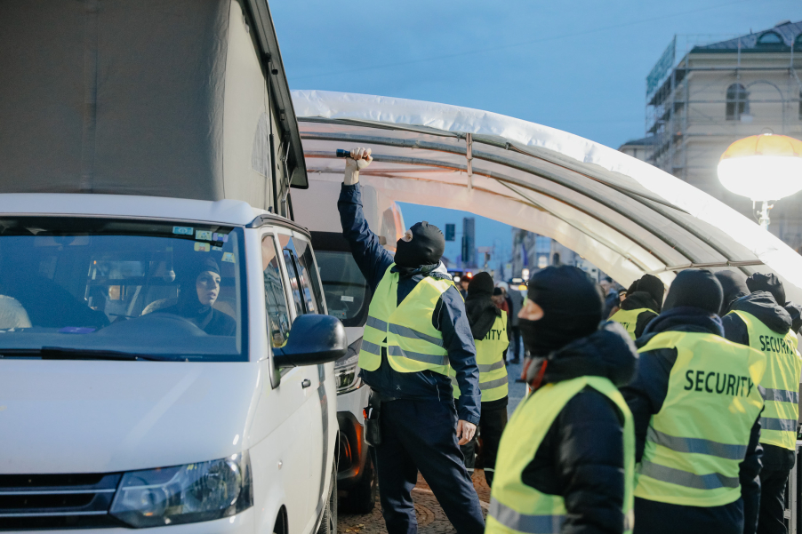 Vermummte Sicherheitskräfte stehen an einem weißen Transporter, eine Sicherheitsperson leuchtet mit einer Taschenlampe in das aufgestellte Hochdach des Transporters.