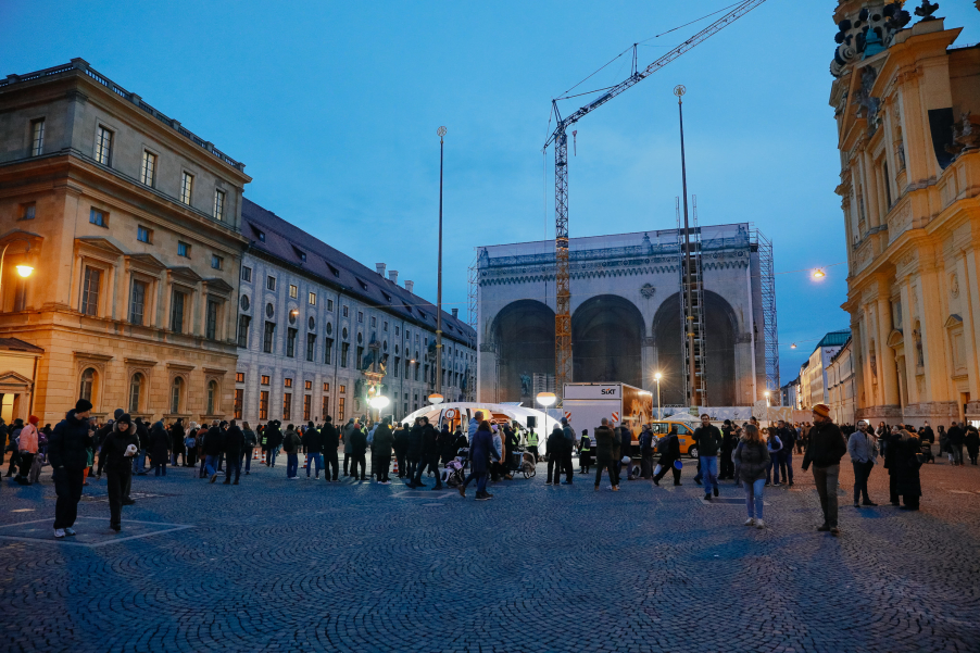 Eine temporäre Kontrollstelle mit Zeltdach steht in der Dämmerung auf einem Platz vor einem historischen Gebäude, der Platz ist mit vier hellen Lampen ausgeleuchtet. Viele Menschen betrachten die Szene.