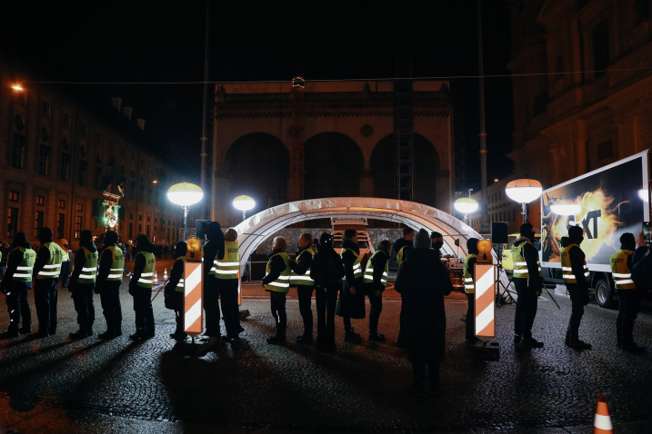 Eine temporäre Kontrollstelle mit Zeltdach steht auf einem Platz vor einem historischen Gebäude. Eine Reihe von Sicherheitskräften steht vor der nächtlichen Szenerie, diese ist mit vier hellen Lampen ausleuchtet.