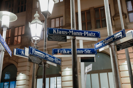 View of several street signs bearing the Mann family name attached to various street lamps. In the background, a historic building facade.