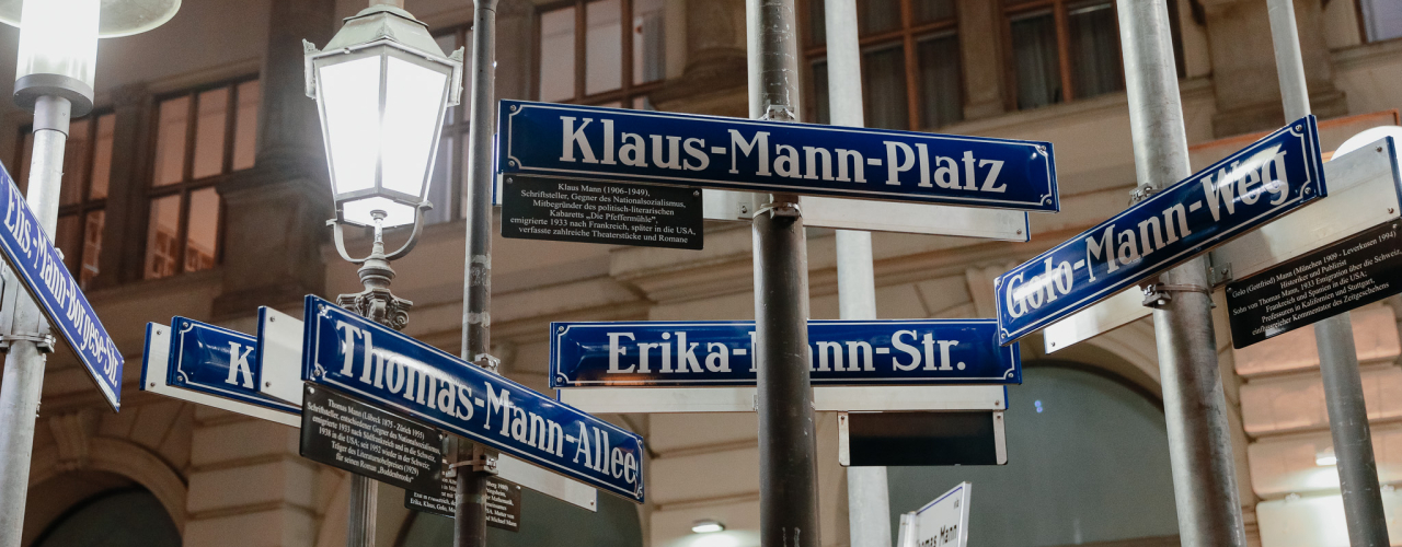 View of several street signs bearing the Mann family name attached to various street lamps. In the background, a historic building facade.