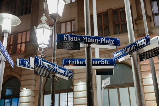 Blick auf einige Straßenschilder mit Namen der Familie Mann an unterschiedlichen Straßenlaternen befestigt. Im Hintergrund eine historische Häuserfassade.