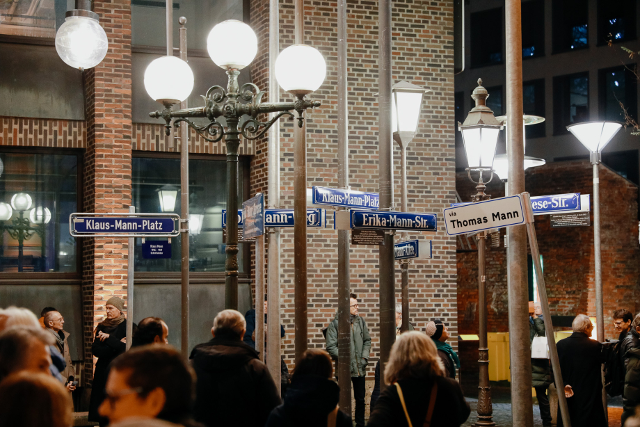 Several people are standing in front of a brick facade between various bright, partly historic street lamps. Street signs with different names of the Mann family are attached to the street lamps.