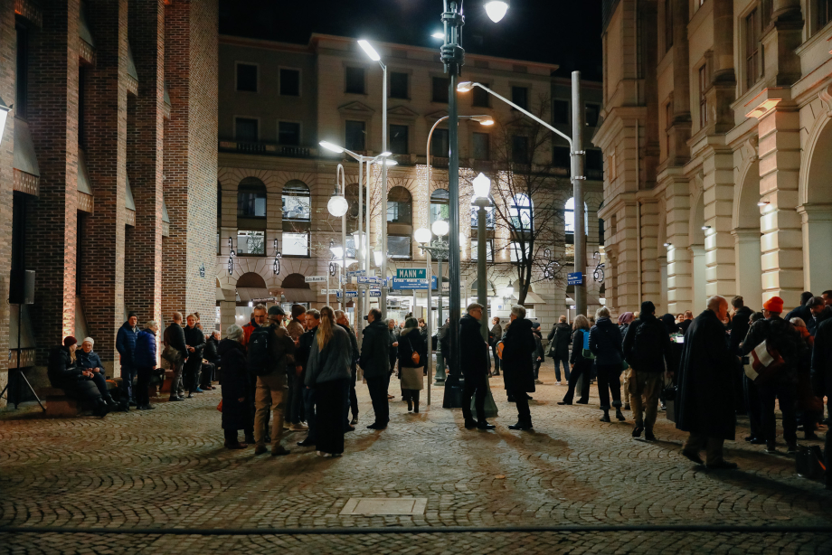 Many people are standing between historic building facades in a square lit by several different street lamps. Various street signs bearing the name of the Mann family are attached to these lamps.