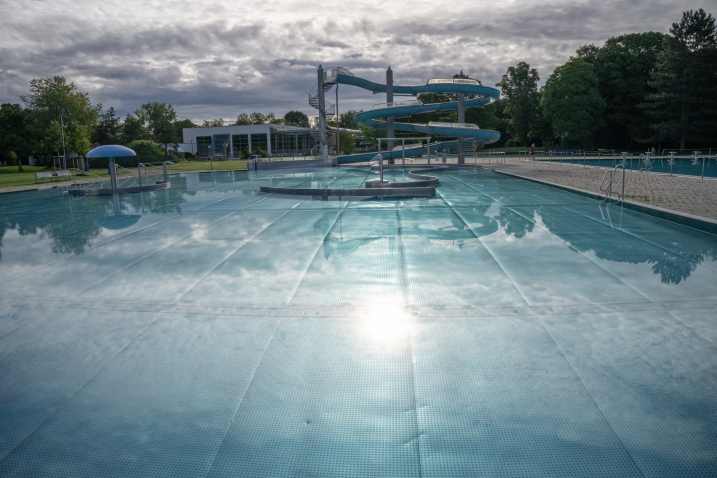 Leeres Becken im Freibad mit geschlängelter Rutsche, im Hintergrund Bäume und Wolkenhimmel.
