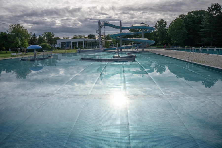 Leeres Becken im Freibad mit geschlängelter Rutsche, im Hintergrund Bäume und Wolkenhimmel.