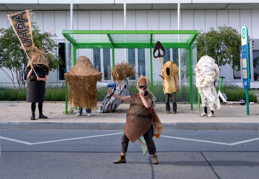 Picture of an artistic performance: Several people are wearing sculptural costumes made of natural or nature-like materials. One person in the foreground is speaking into a megaphone, while the others are standing scattered in front of and inside the bus stop. The scene looks like a staged public performance.