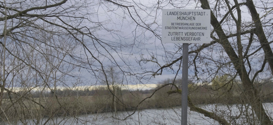 Schild an einem Fluss mit kahlen Bäumen: „Landeshauptstadt München – Betriebsanlage der Stadtentwässerungswerke – Zutritt verboten, Lebensgefahr."