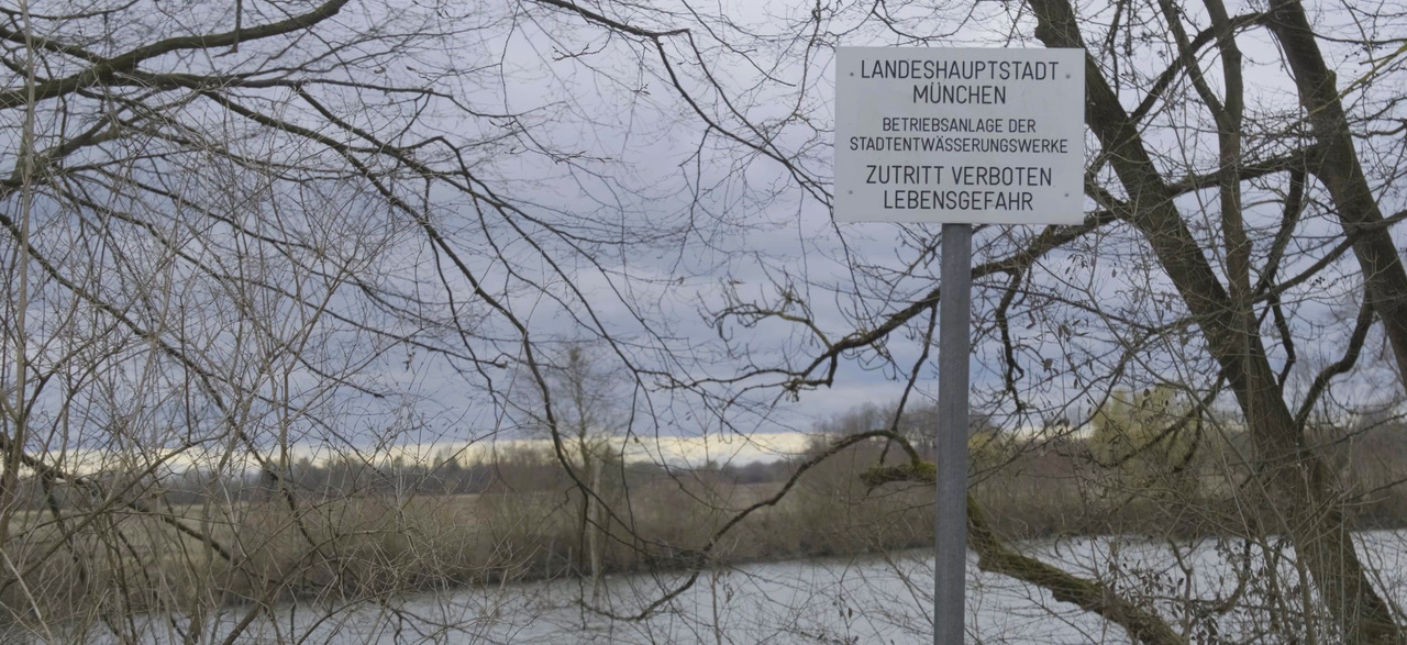 Schild an einem Fluss mit kahlen Bäumen: „Landeshauptstadt München – Betriebsanlage der Stadtentwässerungswerke – Zutritt verboten, Lebensgefahr."