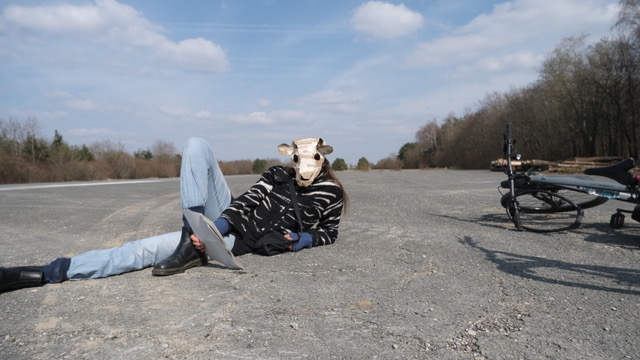 Eine Person mit Schafmaske liegt entspannt auf einer leeren Straße, neben ihr liegt ein Fahrrad.