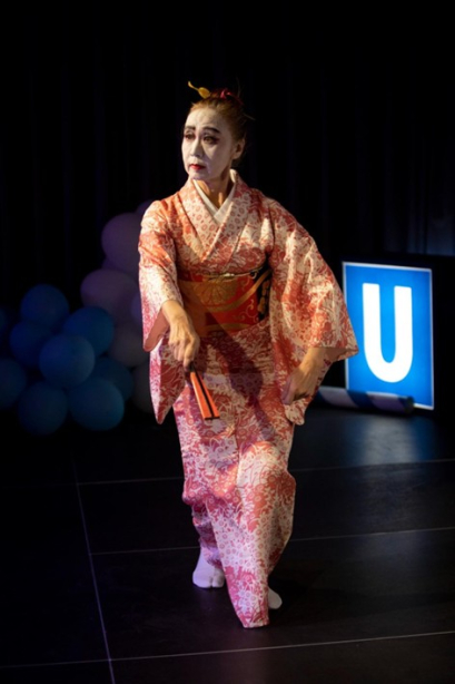 Person in rosa Kimono mit weißer Gesichtsschminke tanzt auf einer Bühne, im Hintergrund ein U-Bahn-Schild und Luftballons.