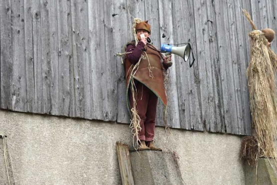 Vier kostümierte Figuren aus Stroh und Naturmaterialien stehen auf Betonpfeilern vor einer Holzscheune, eine davon ist eine echte Person mit einem Megaphon in der Hand.