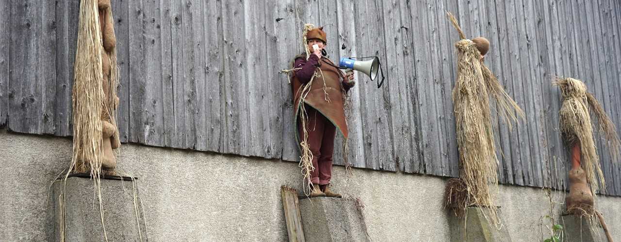 Vier kostümierte Figuren aus Stroh und Naturmaterialien stehen auf Betonpfeilern vor einer Holzscheune, eine davon ist eine echte Person mit einem Megaphon in der Hand.
