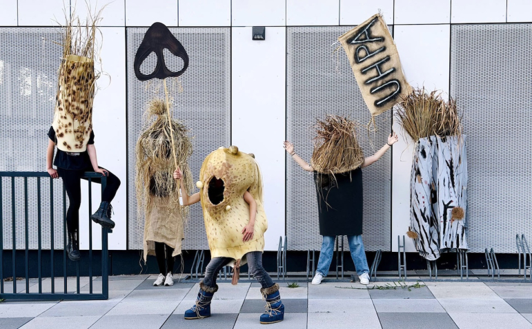 Five people are standing in front of the facade of a modern building, wearing large, sculptural costumes made of natural-looking materials. The costumes resemble plants, straw, bark, and a yellowish, porous object. Two people are holding signs with a black symbol and the letters “UHPA.” One person in a yellow costume is stepping forward, while another is sitting on a metal railing.