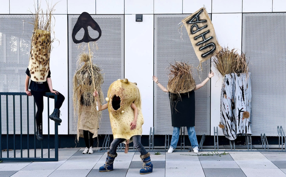 Five people are standing in front of the facade of a modern building, wearing large, sculptural costumes made of natural-looking materials. The costumes resemble plants, straw, bark, and a yellowish, porous object. Two people are holding signs with a black symbol and the letters “UHPA.” One person in a yellow costume is stepping forward, while another is sitting on a metal railing.