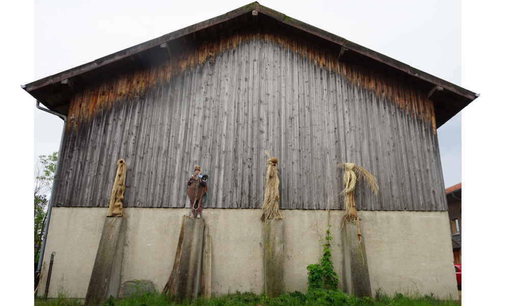 Vier kostümierte Figuren aus Stroh und Naturmaterialien stehen auf Betonpfeilern vor einer Holzscheune, eine davon ist eine echte Person mit einem Megaphon in der Hand.