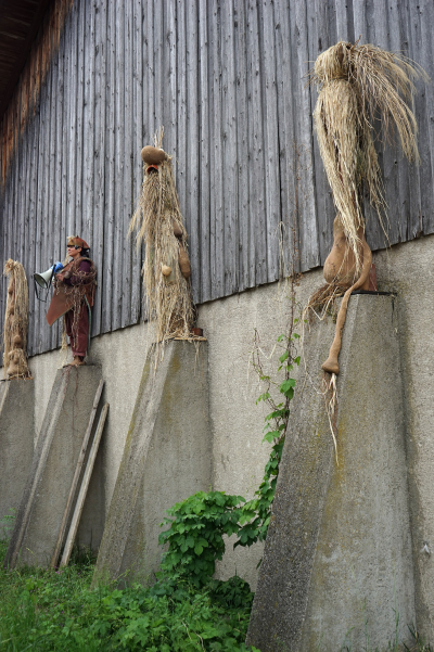 Vier kostümierte Figuren aus Stroh und Naturmaterialien stehen auf Betonpfeilern vor einer Holzscheune, eine davon ist eine echte Person mit einem Megaphon in der Hand.
