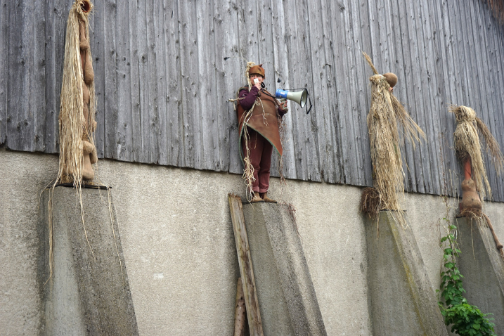 Vier kostümierte Figuren aus Stroh und Naturmaterialien stehen auf Betonpfeilern vor einer Holzscheune, eine davon ist eine echte Person mit einem Megaphon in der Hand.