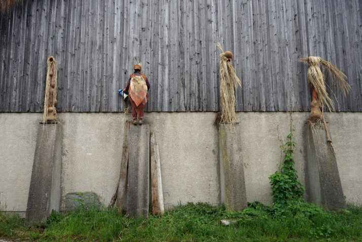 Vier kostümierte Figuren aus Stroh und Naturmaterialien stehen auf Betonpfeilern vor einer Holzscheune, eine davon ist eine echte Person mit dem Rücken zur Kamera.