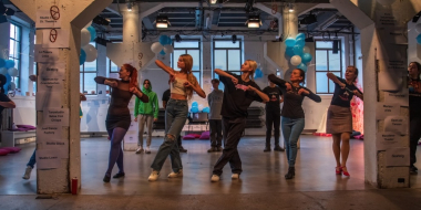 Eight people perform a choreographed dance together in a decorated event hall. They stand in a line, moving in sync, while other participants watch or join in from the background. Blue and white balloons and directional signs on the pillars give the space the atmosphere of a dance or cultural event.
