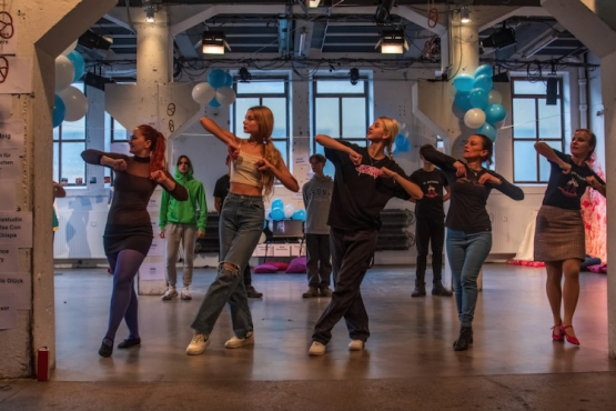 Acht Personen führen in einer dekorierten Veranstaltungshalle gemeinsam eine Tanzchoreografie auf. Sie stehen in einer Reihe und bewegen sich synchron, während weitere Teilnehmende im Hintergrund zusehen oder mitmachen. Blau-weiße Luftballons und Wegweiser an den Säulen verleihen dem Raum den Charakter einer Tanz- oder Kulturveranstaltung.
