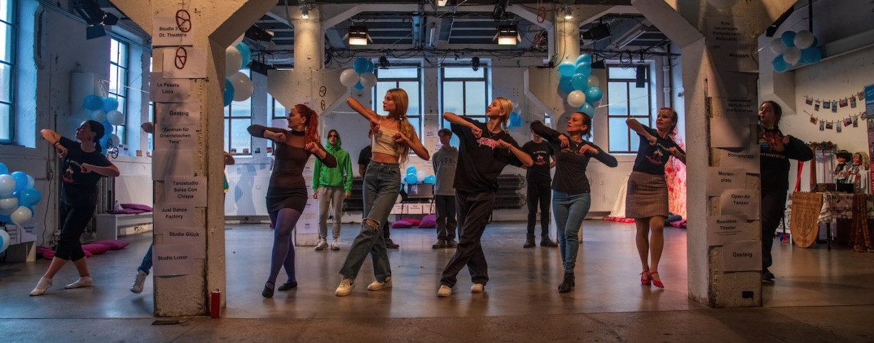 Acht Personen führen in einer dekorierten Veranstaltungshalle gemeinsam eine Tanzchoreografie auf. Sie stehen in einer Reihe und bewegen sich synchron, während weitere Teilnehmende im Hintergrund zusehen oder mitmachen. Blau-weiße Luftballons und Wegweiser an den Säulen verleihen dem Raum den Charakter einer Tanz- oder Kulturveranstaltung.