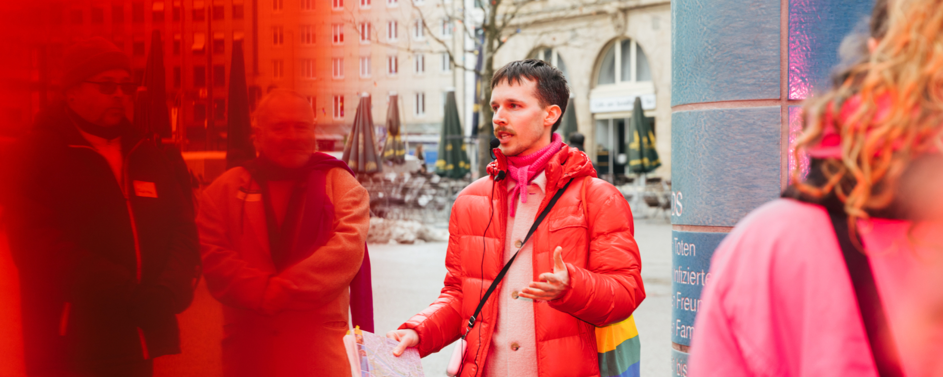 Der Gay Guide Anian Halder trägt eine rote Jacke. Er steht mittig im Bild. Rechts neben ihm ist eine blaue Säule zu sehen. Die linke Bildseite ist rot-transparent überlagert. Um ihn stehen Personen.
