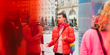 Gay guide Anian Halder is wearing a red jacket. He is standing in the center of the picture. To his right is a blue column. The left side of the picture is overlaid with a red transparent layer. There are people standing around him.