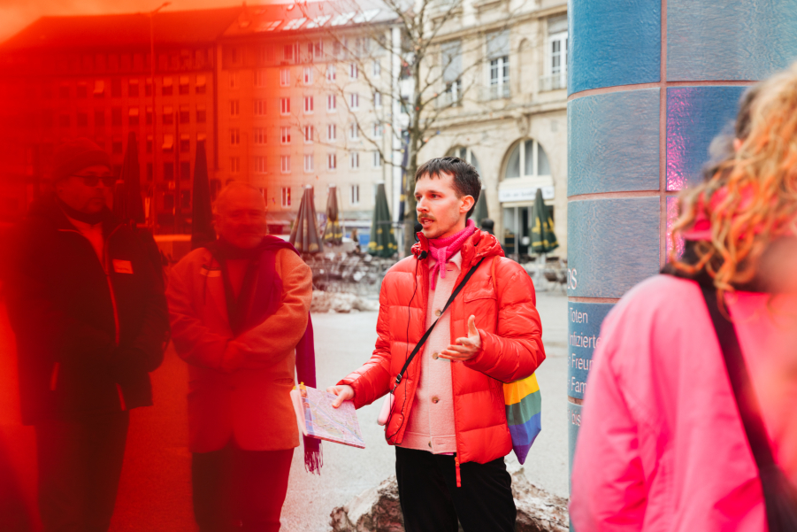 A person wearing a red down jacket and carrying a rainbow-colored bag stands in front of a tiled column in a city square and speaks to a group of people.