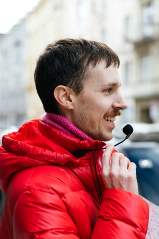 Gay guide Anian Halder can be seen in profile. He is wearing a red jacket and a pink scarf. He is touching a microphone attached to his jacket with his hand.