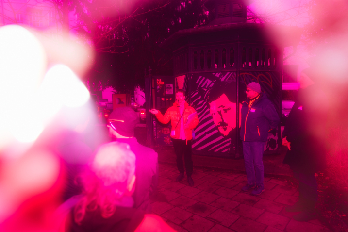 A blurred image shows the outlines of people. In the center stands Gay Guide Anian Halder in front of a toilet block. He raises his left hand. To his right stands a guest on the city tour. The image is overlaid with a red transparent layer.
