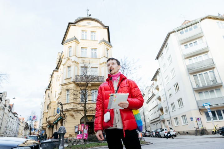In the center of the picture is Gay Guide Anian Halder. He is standing in front of a yellow building. He is holding a booklet in his hand.