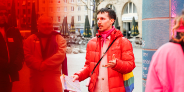 Eine Person in roter Daunenjacke mit einem Stofftasche in Regenbogenfarben steht vor einer gekachelten Säule auf einem Stadtplatz und spricht zu einer Gruppe von Menschen.
