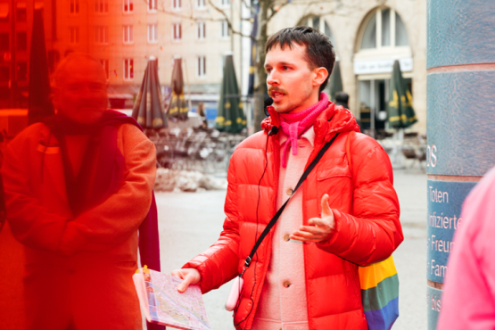 A person wearing a red down jacket and carrying a rainbow-colored bag stands in front of a tiled column in a city square and speaks to a group of people.