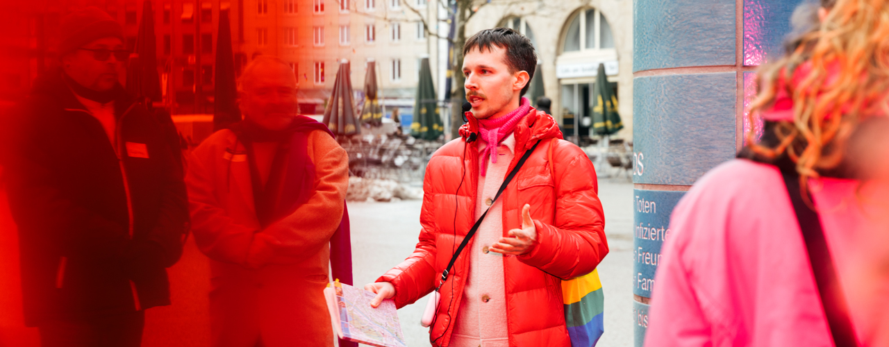 A person wearing a red down jacket and carrying a rainbow-colored bag stands in front of a tiled column in a city square and speaks to a group of people.