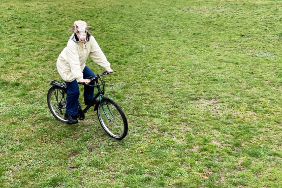Person mit Schafsmaske fährt Fahrrad über eine Wiese.