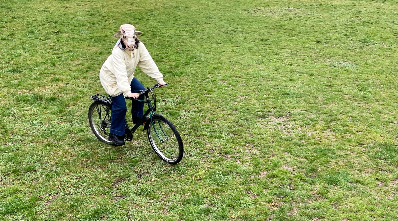 Person mit Schafsmaske fährt Fahrrad über eine Wiese.