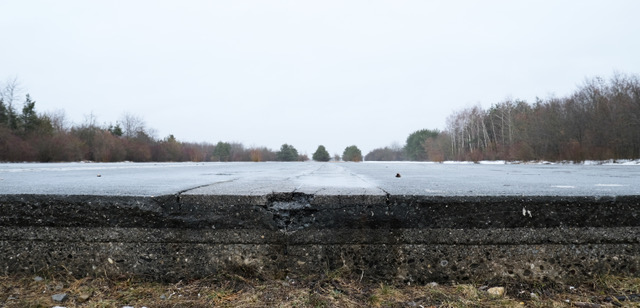 Verlassene, rissige Asphaltfläche im Winter, umgeben von kahlen Bäumen und grauem Himmel.