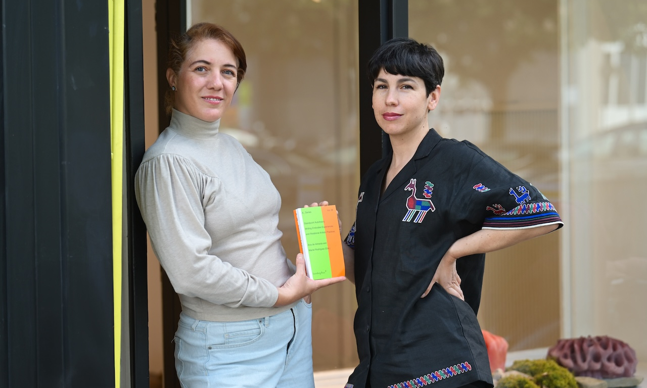 Zwei Frauen stehen nebeneinander vor einer großen Glasfront. Die Frau links trägt einen hellgrauen Rollkragenpullover und helle Jeans und hält ein buntes Buch in den Händen. Die Frau rechts trägt eine schwarze Bluse mit farbigen Stickmustern und hat eine Hand in die Hüfte gestützt. Beide blicken in die Kamera. Im Hintergrund sind durch die Fensterscheibe unscharfe Dekorationen und Reflexionen zu sehen.