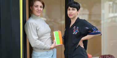Two women are standing side by side in front of a large glass window. The woman on the left is wearing a light gray turtleneck and light-colored jeans, and is holding a colorful book in her hands. The woman on the right is wearing a black blouse with colorful embroidery and has one hand on her hip. Both are looking at the camera. In the background, blurred decorations and reflections can be seen through the windowpane.