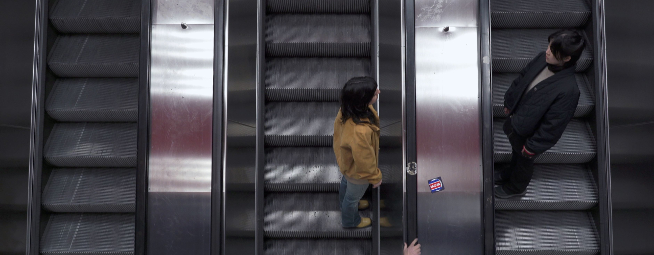 Zwei Personen fahren auf parallelen Rolltreppen in entgegengesetzte Richtungen. Die Szene ist von oben aufgenommen, mit metallischen Oberflächen und dunklen Stufen, wodurch eine symmetrische, urbane Atmosphäre entsteht.