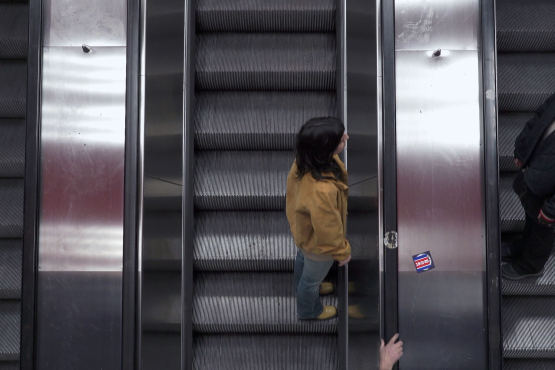 Zwei Personen fahren auf parallelen Rolltreppen in entgegengesetzte Richtungen. Die Szene ist von oben aufgenommen, mit metallischen Oberflächen und dunklen Stufen, wodurch eine symmetrische, urbane Atmosphäre entsteht.