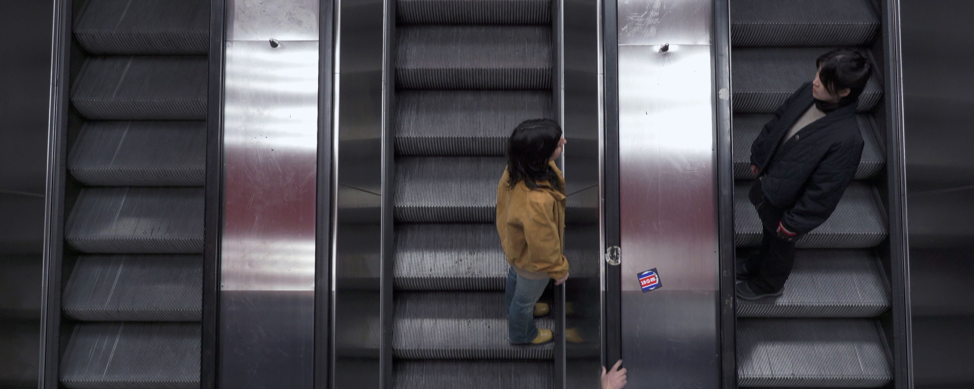 Zwei Personen fahren auf parallelen Rolltreppen in entgegengesetzte Richtungen. Die Szene ist von oben aufgenommen, mit metallischen Oberflächen und dunklen Stufen, wodurch eine symmetrische, urbane Atmosphäre entsteht.