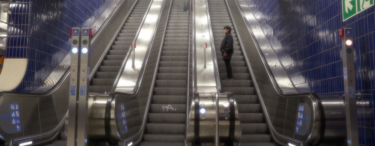 Breite Rolltreppenanlage in einer U-Bahn-Halle mit dunkelblauen Fliesenwänden. Zwei Personen stehen auf verschiedenen Rolltreppen und wenden sich aneinander zu.